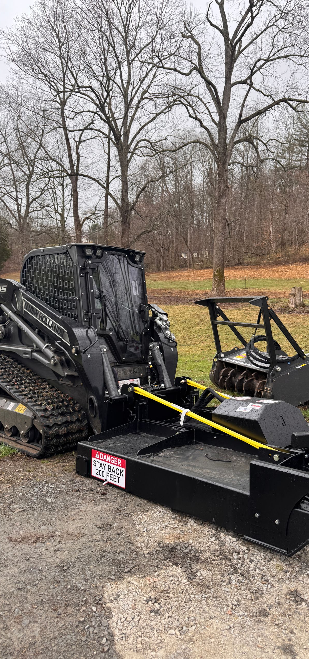 Black riding lawn mower on a trailer in a gravel lot with bare trees in the background