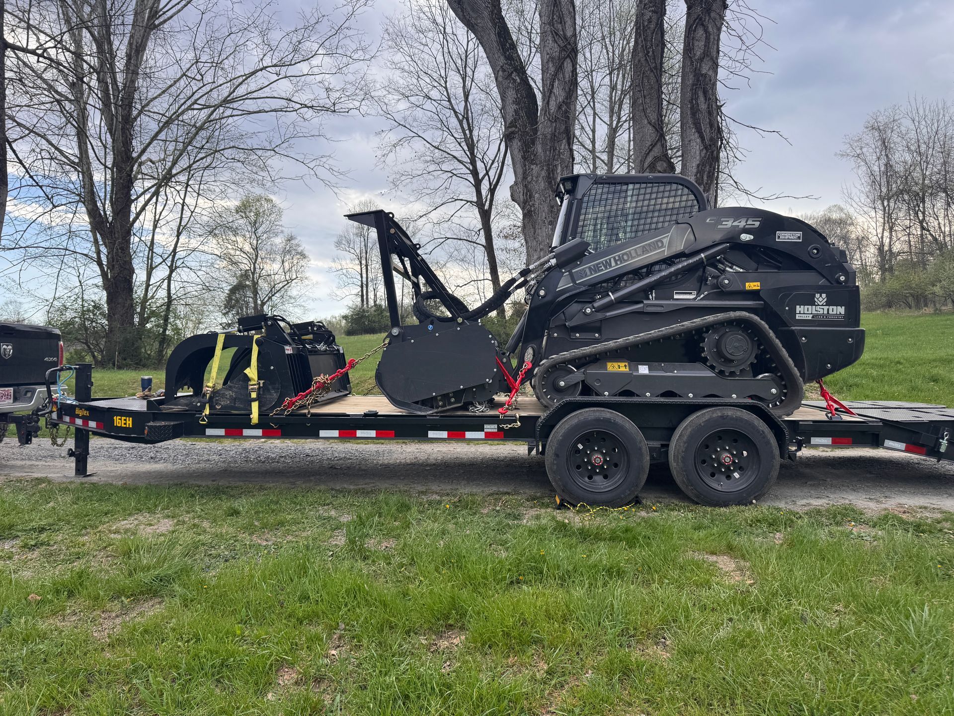 Black construction equipment on a trailer in a grassy roadside area with bare trees in the background