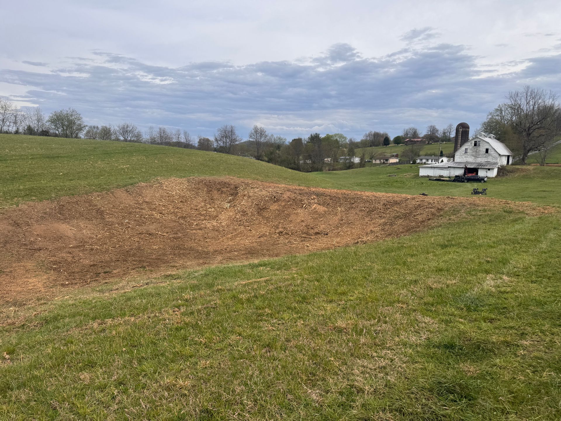 Grassy field with a large brown dirt mound and a white house in the distance under a cloudy sky