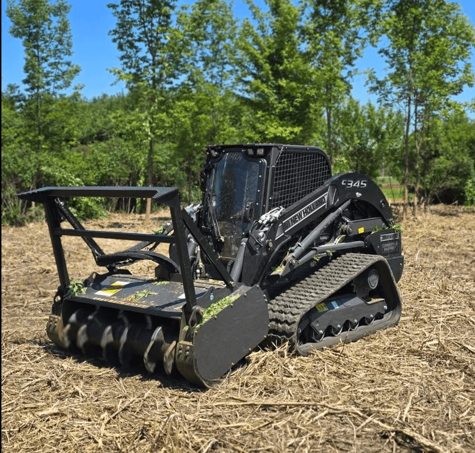 Black tracked compact loader with grapple attachment on a wood-chipped site under green trees