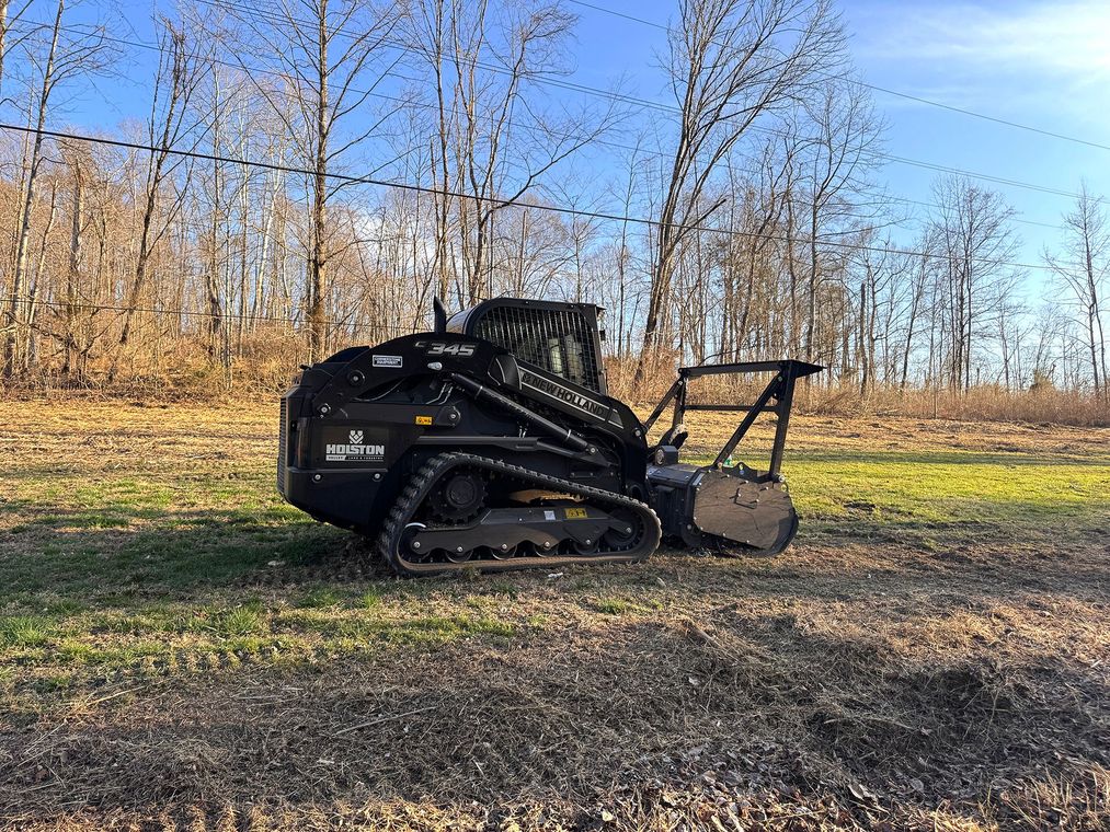 Black tracked skid-steer loader parked on gravel in a leafless wooded area under a blue sky.