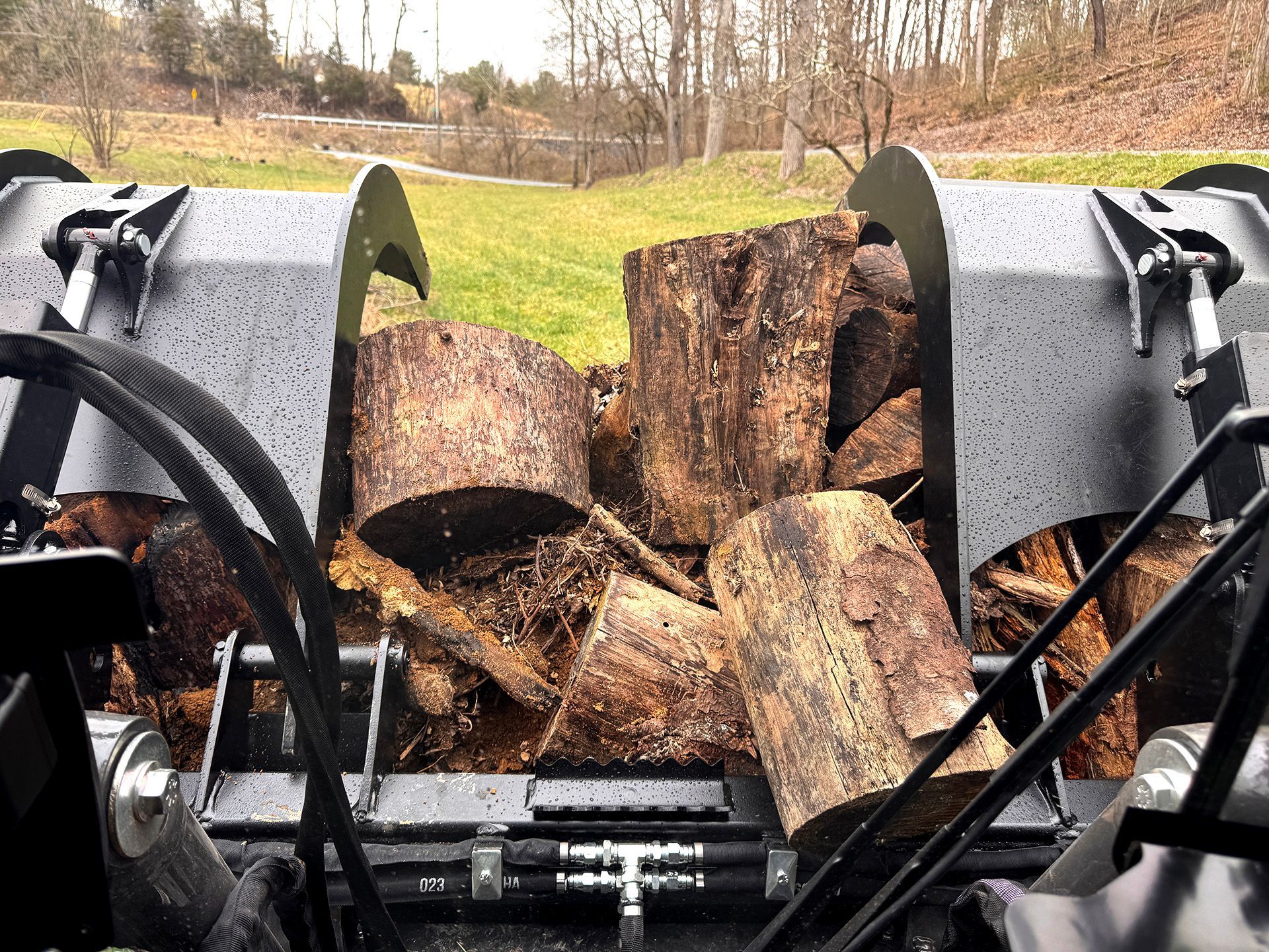 Two machine arms hold split logs in a wood chipper outdoors on a grassy yard.