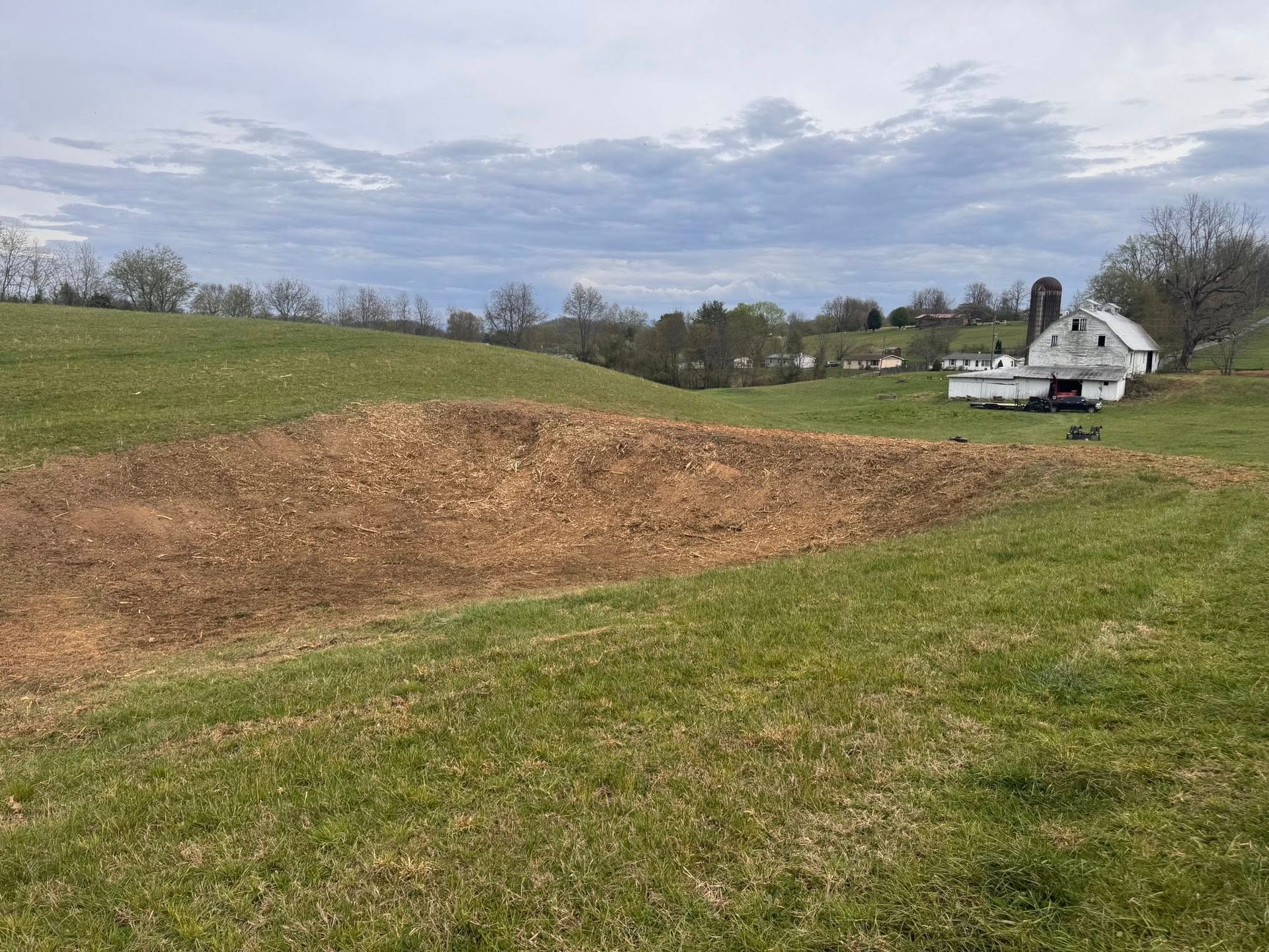 Grassy field with a large brown dirt mound under a cloudy sky, with houses in the distance