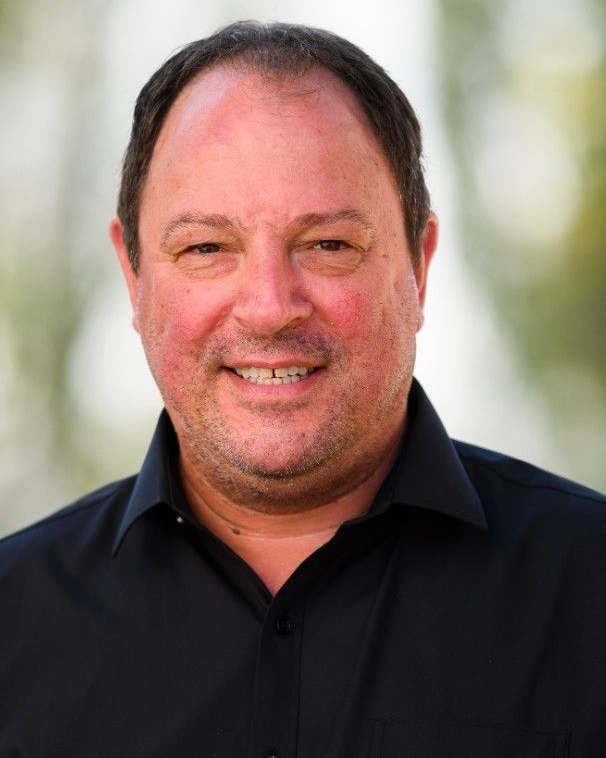 Man with short dark hair, smiling, wearing a black shirt, set outdoors with blurred greenery.