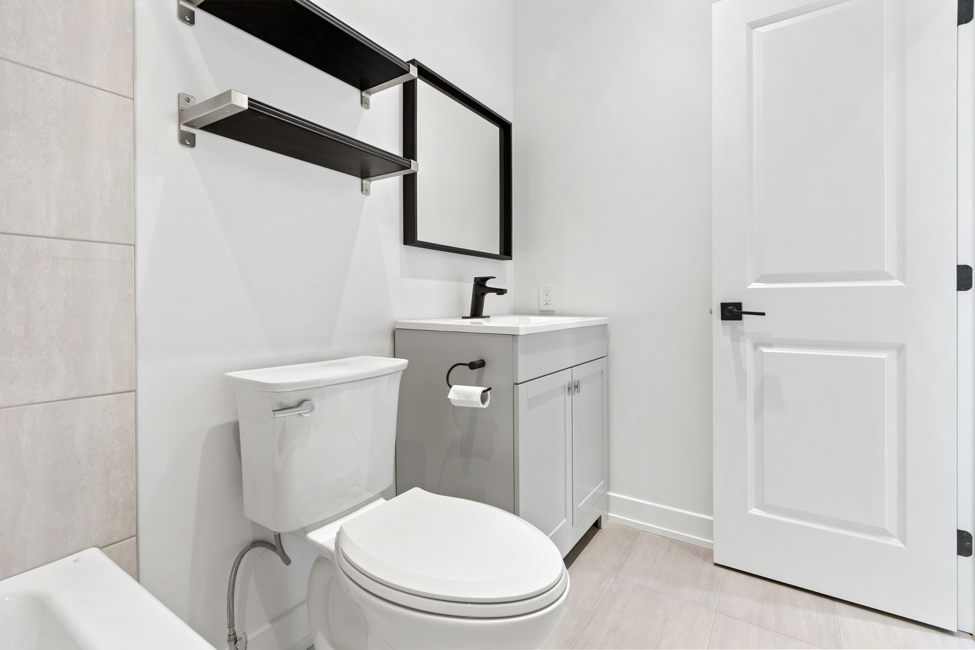 Modern white bathroom with toilet, vanity, mirror, shelves, and closed door.