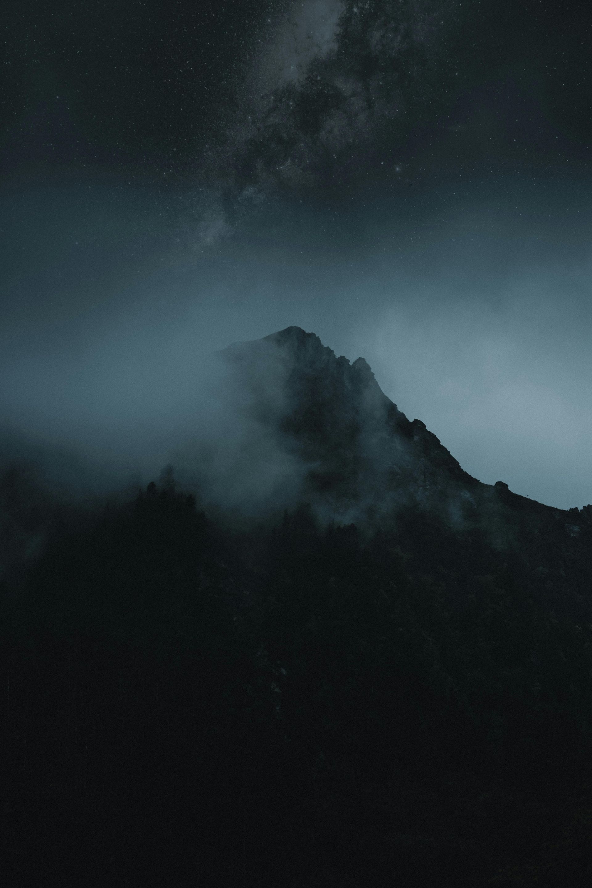A mountain stands tall in the darkness with a light illuminating the space and sky behind it.