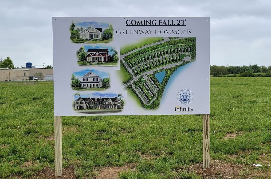 A construction sign in a field displays a site plan and house illustrations for the upcoming