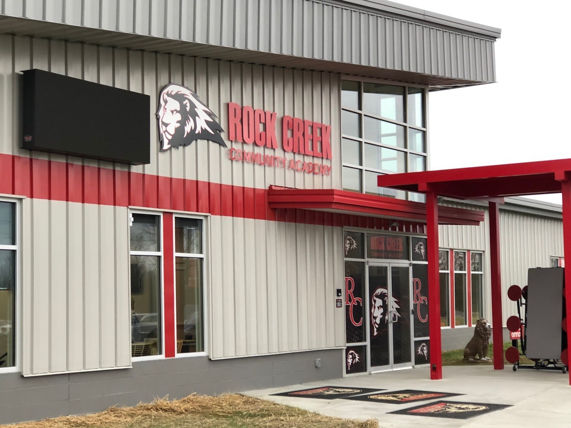 Exterior view of Rock Creek Community Academy with a lion mascot logo, red accents, and a covered entryway.