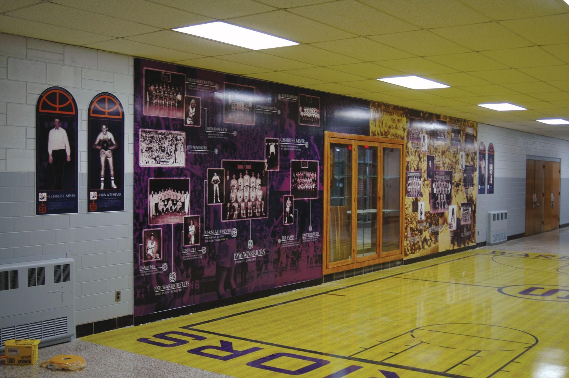 A gymnasium hallway featuring sports memorabilia wall murals, framed portraits, and a yellow basketball court floor.