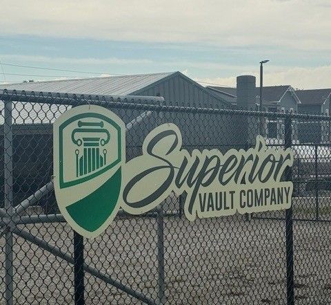 A sign for Superior Vault Company featuring a green column logo on a chain-link fence in front of a building.