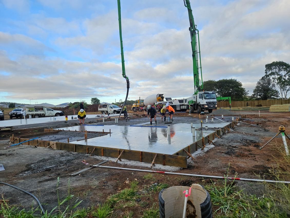 Construction site with concrete slab being poured, workers, and a large crane pump in the background.
