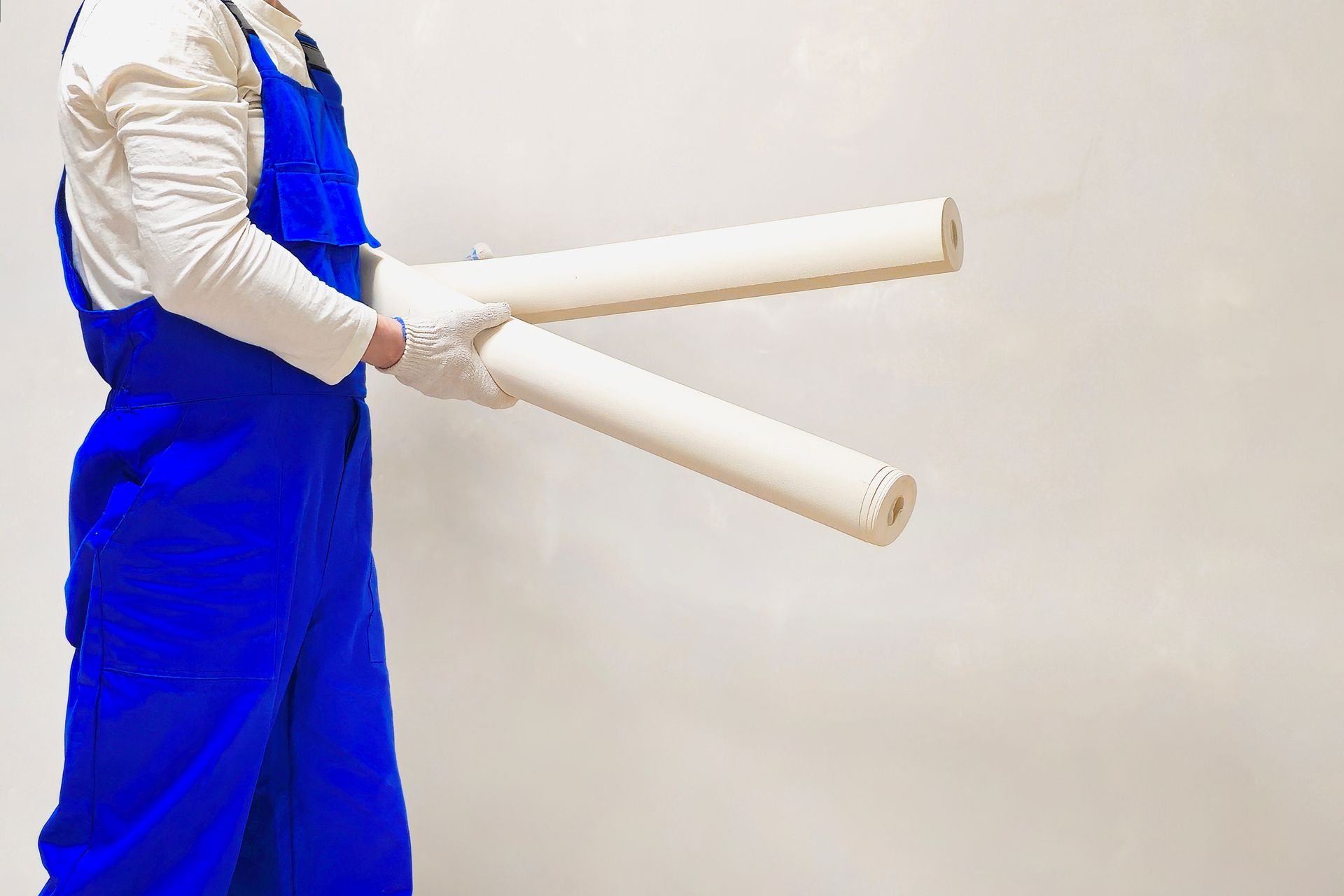 Person in blue overalls holding two rolled papers against a white wall.