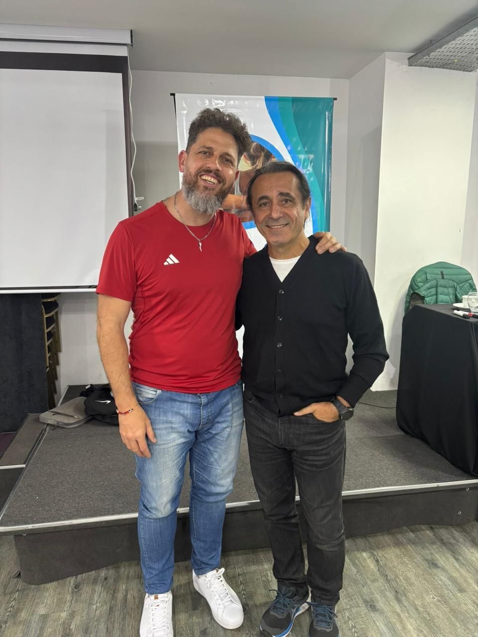 Dos hombres sonriendo y de pie juntos en un escenario en un lugar cerrado, uno con camisa roja y vaqueros, el otro de negro.