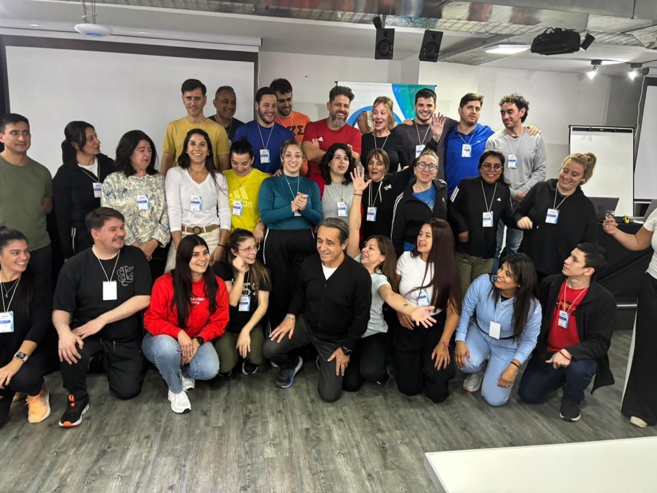 Un grupo numeroso posa junto en el interior, sonriendo en una luminosa habitación que parece una oficina, con suelos grises y paredes blancas.
