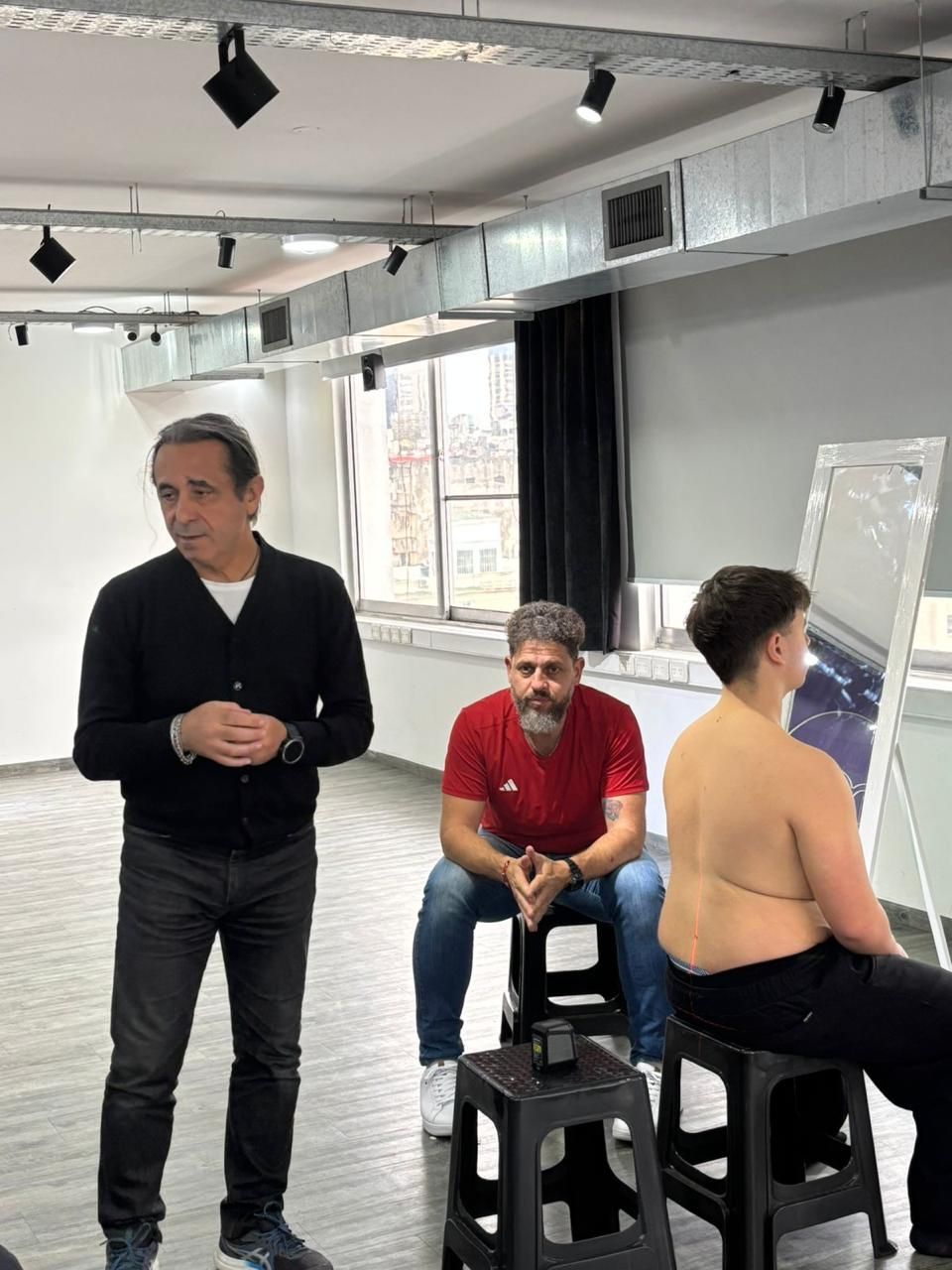 Tres personas en un estudio, una de pie y dos sentadas en taburetes cerca de una pizarra blanca y un caballete.