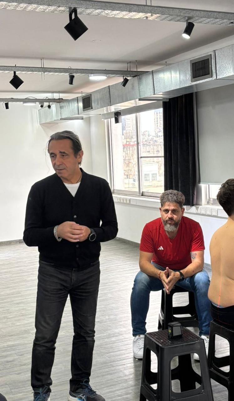 Gente en un estudio luminoso con focos empotrados; un hombre está de pie delante mientras otro está sentado en taburetes apilados.
