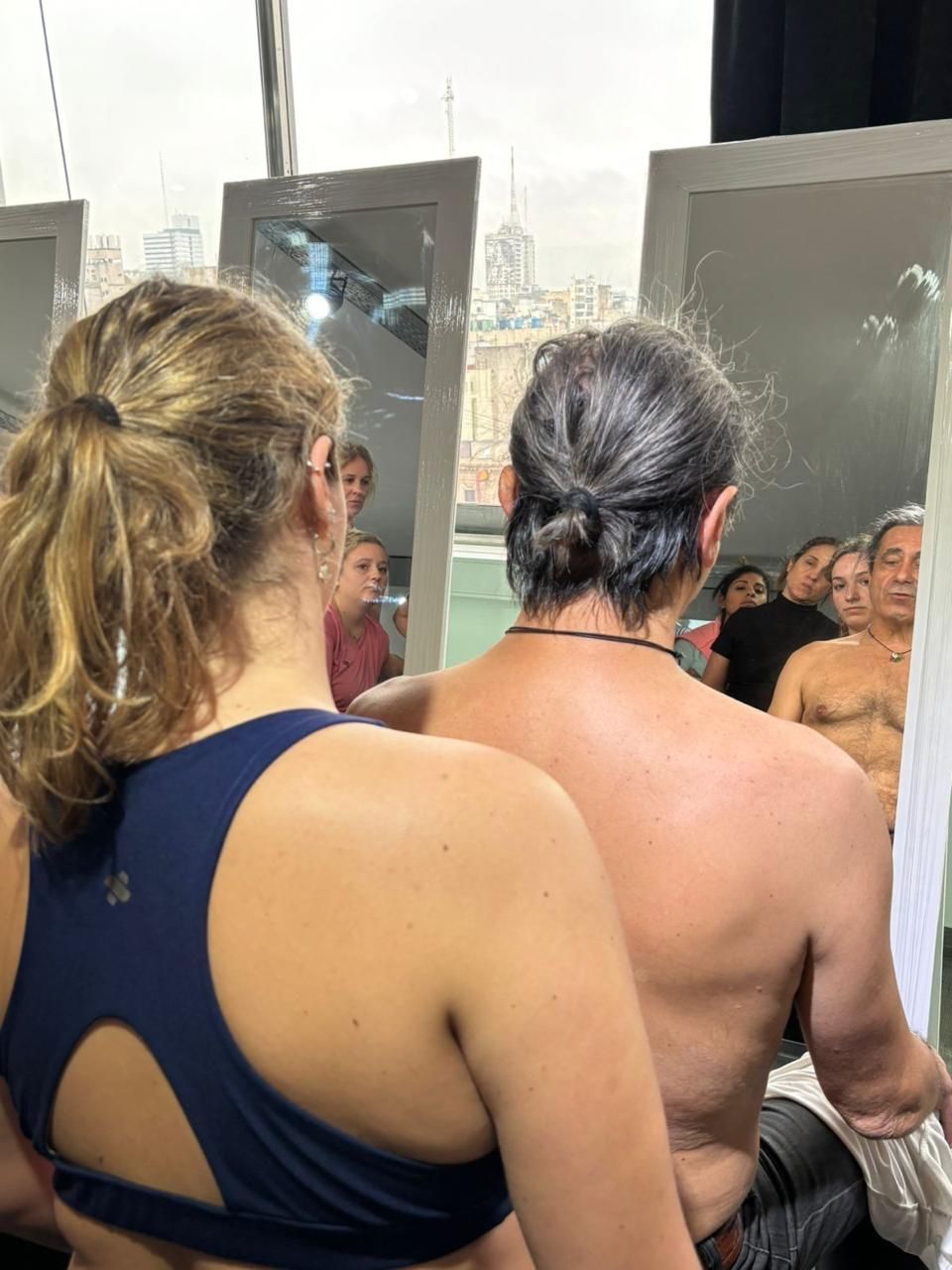 Dos personas con camisetas sin mangas vistas de espaldas, frente a una escena interior concurrida cerca de grandes ventanales.