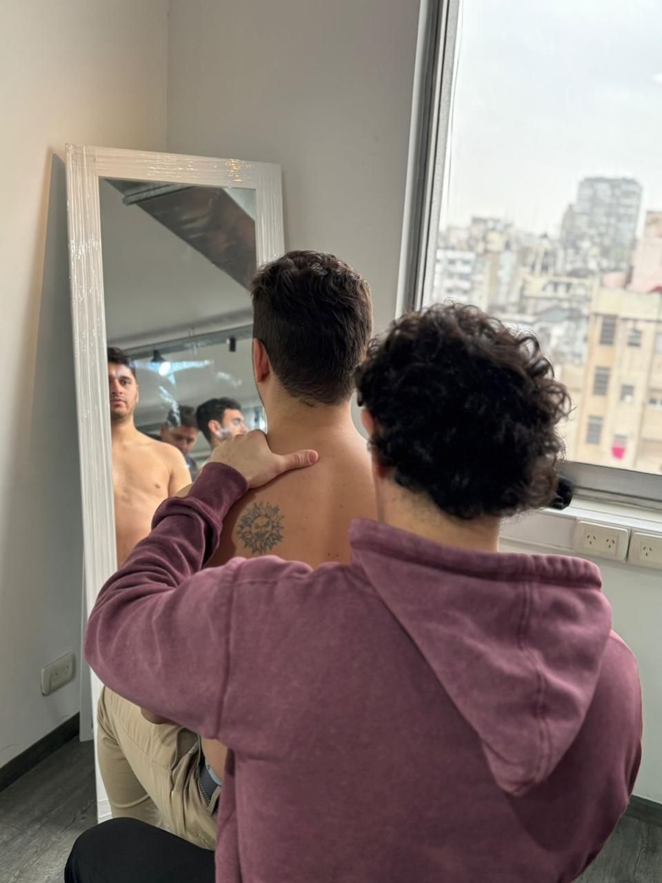 Una persona le está ajustando el cuello de la camisa a otra persona frente a un espejo cerca de una ventana.
