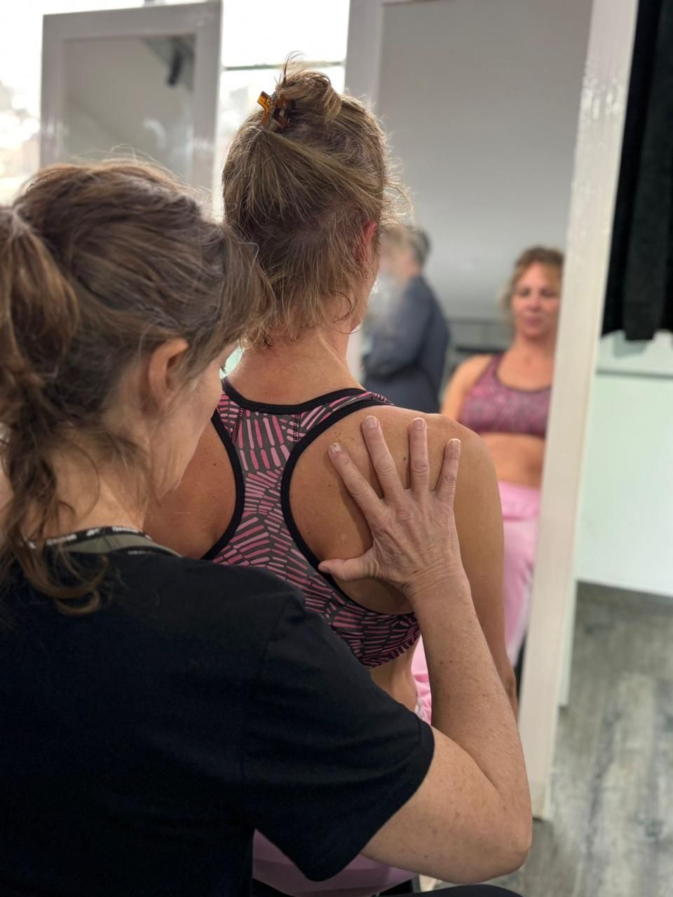Una mujer guía a una niña en un estudio de danza; ambas son vistas desde atrás, con un espejo que refleja su postura.