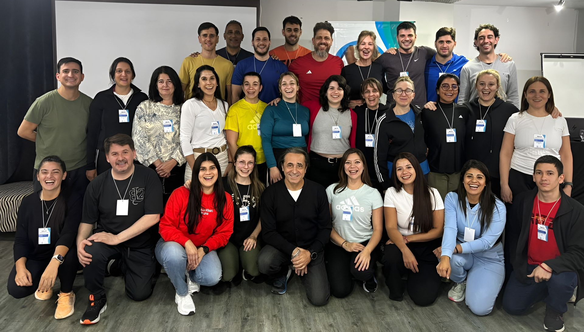 Grupo de 18 personas posando en el interior, en dos filas, sonriendo y vestidas con ropa informal y credenciales de identificación.
