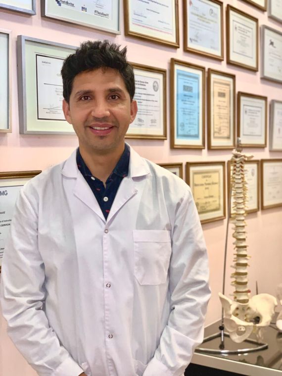 Un médico con bata blanca de laboratorio está de pie en una oficina con certificados enmarcados y un modelo de columna vertebral al fondo.