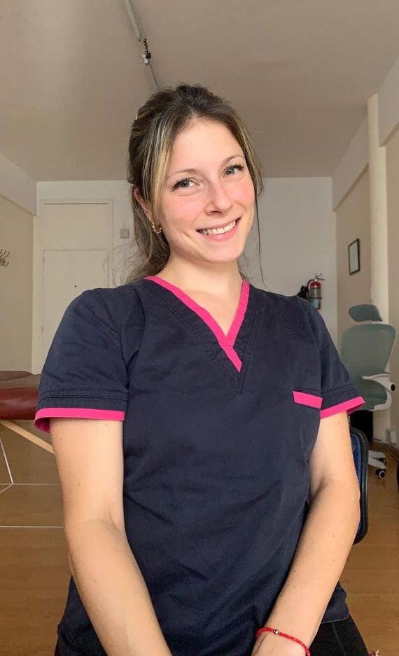 Persona sonriente con uniforme médico azul marino con ribete rosa, de pie en el interior.