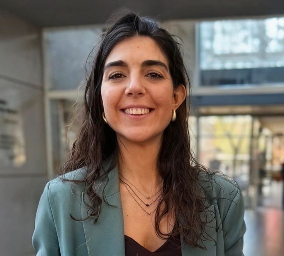 Una persona sonriente con un blazer color turquesa está de pie al aire libre en el patio de un edificio moderno.