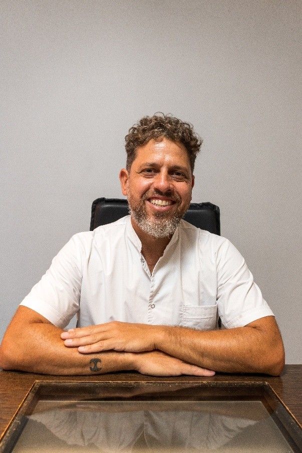 Hombre sonriente con camisa blanca sentado en un escritorio contra una pared gris.