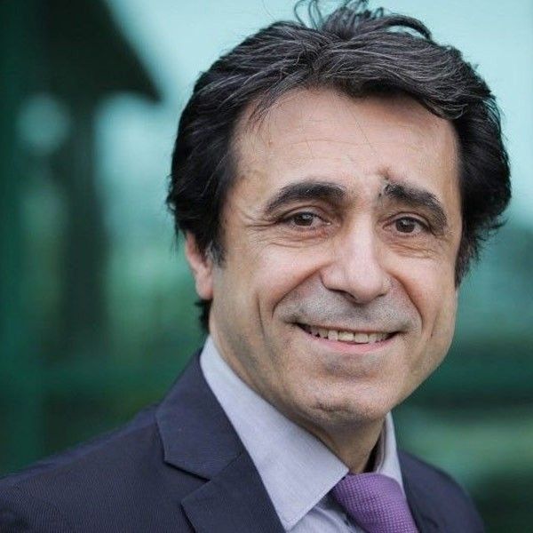 Retrato de un hombre sonriente con traje oscuro y corbata morada sobre un fondo verde difuminado.