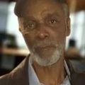 Close-up of an older man with a gray beard, looking at the camera indoors.