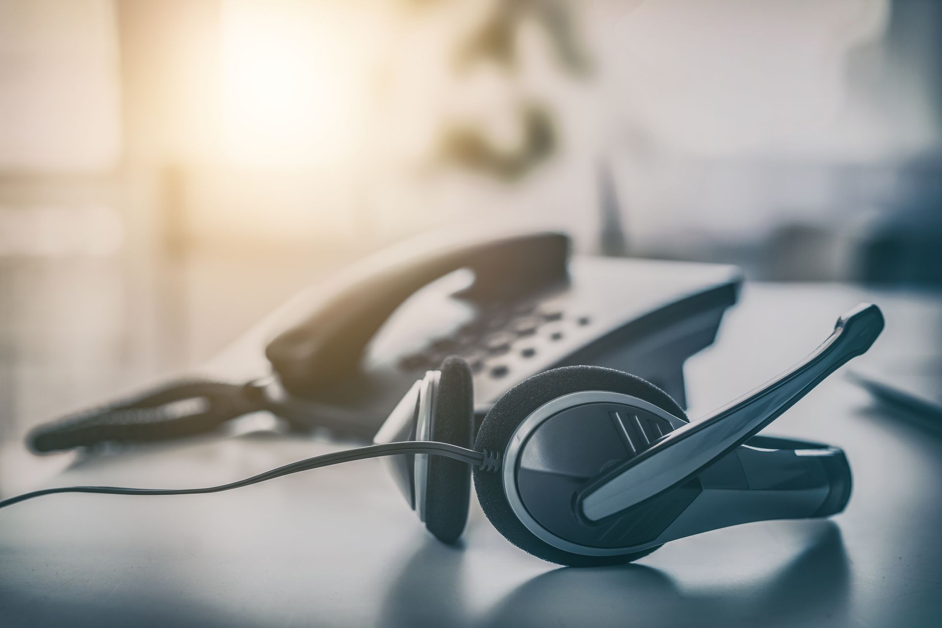 Black office headset beside a desk phone on a blurred tabletop with warm light background