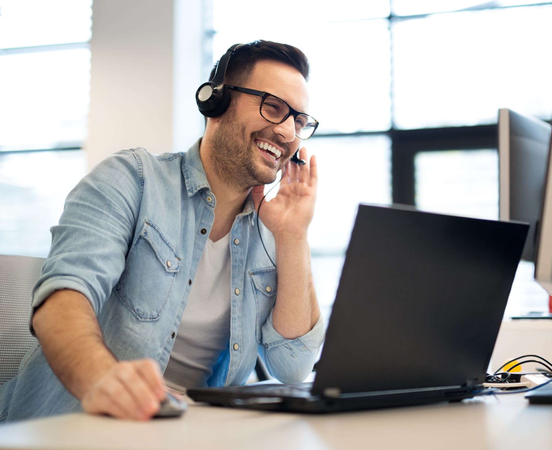 Man wearing headphones and glasses talking on phone at a laptop in a bright office