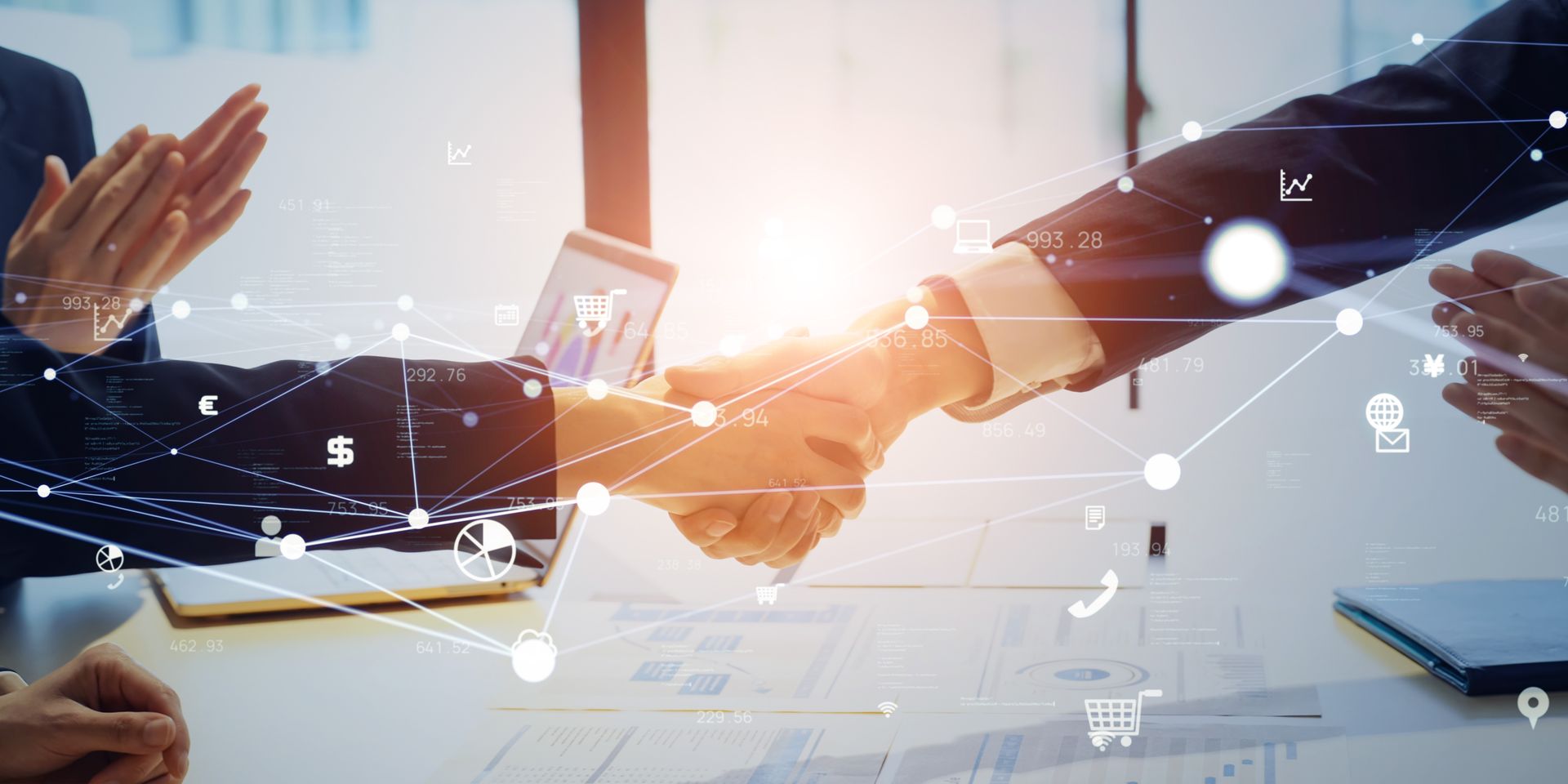 Businesspeople shaking hands across a desk with a laptop and digital network icons overlayed