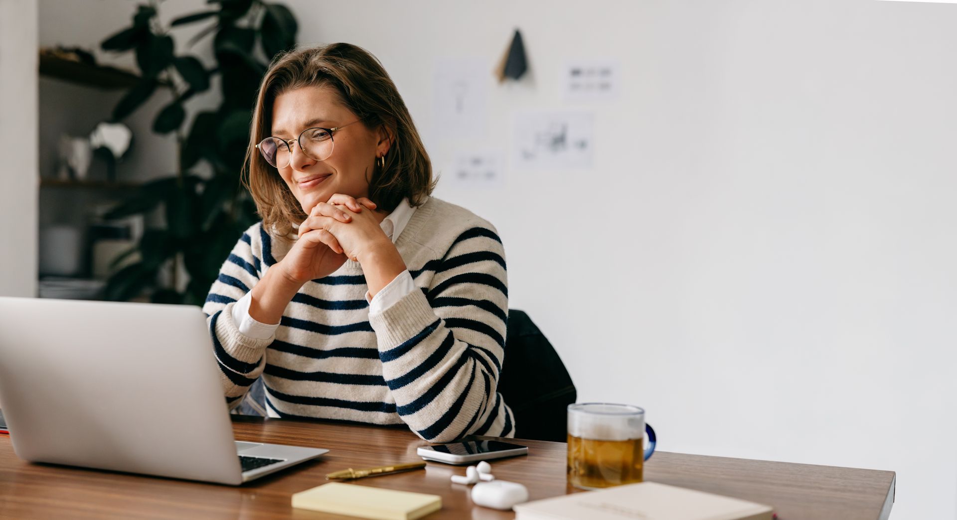 Person smiling beside a laptop at a desk, holding a cup in a bright office setting.