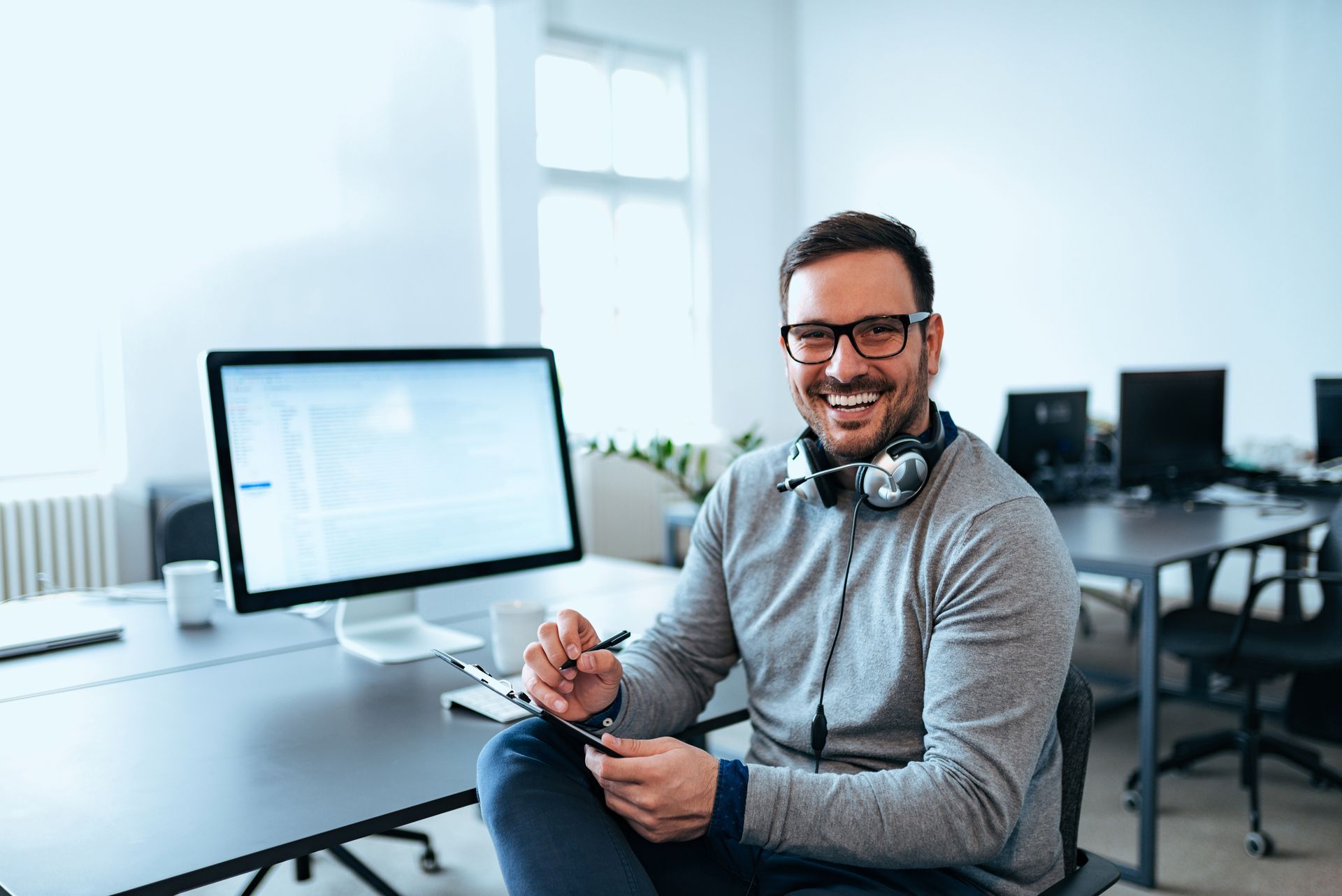Man smiling at a desk in a bright office, holding a stylus beside a computer monitor