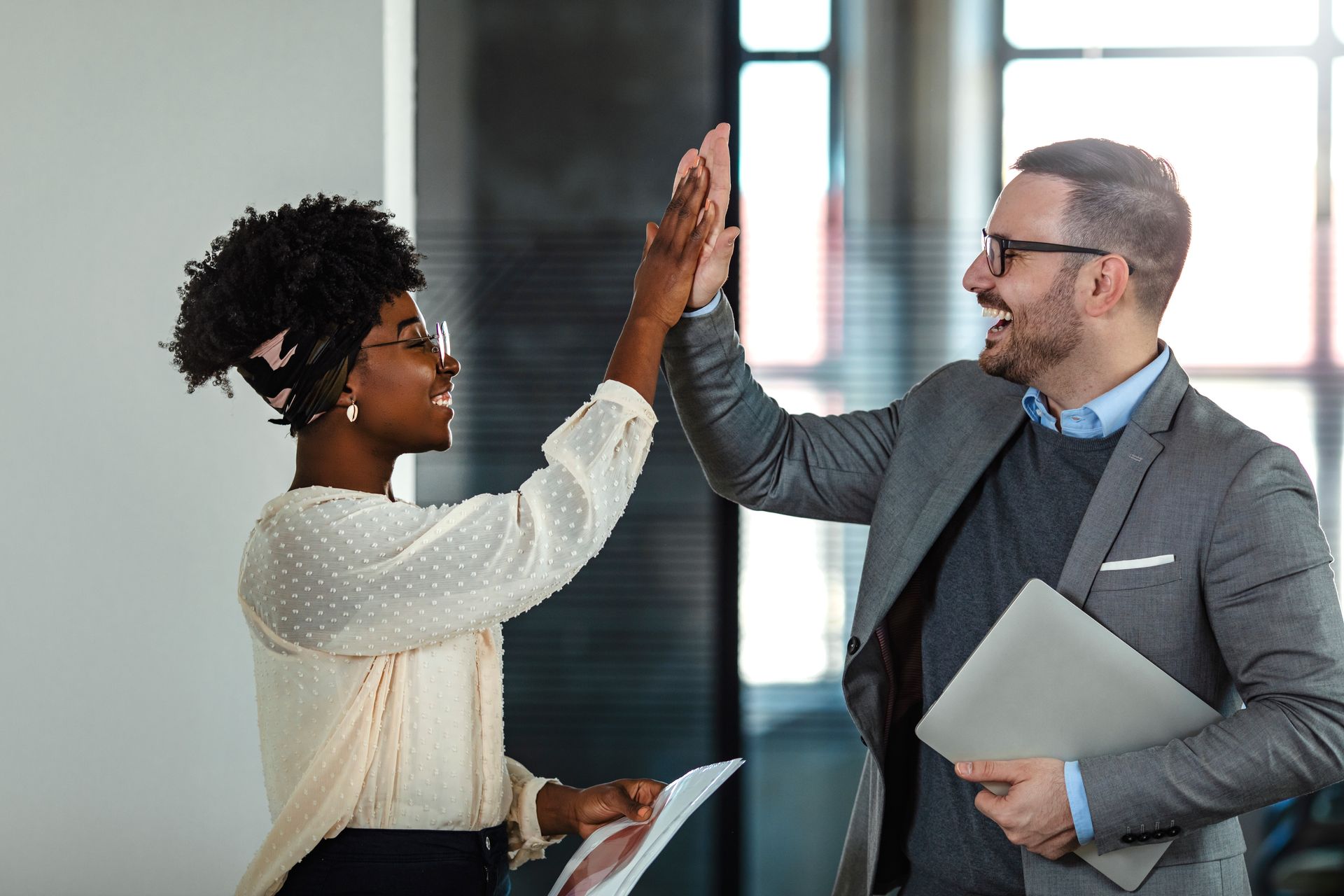 Two coworkers high-five in an office hallway, one holding a folder and the other a tablet.