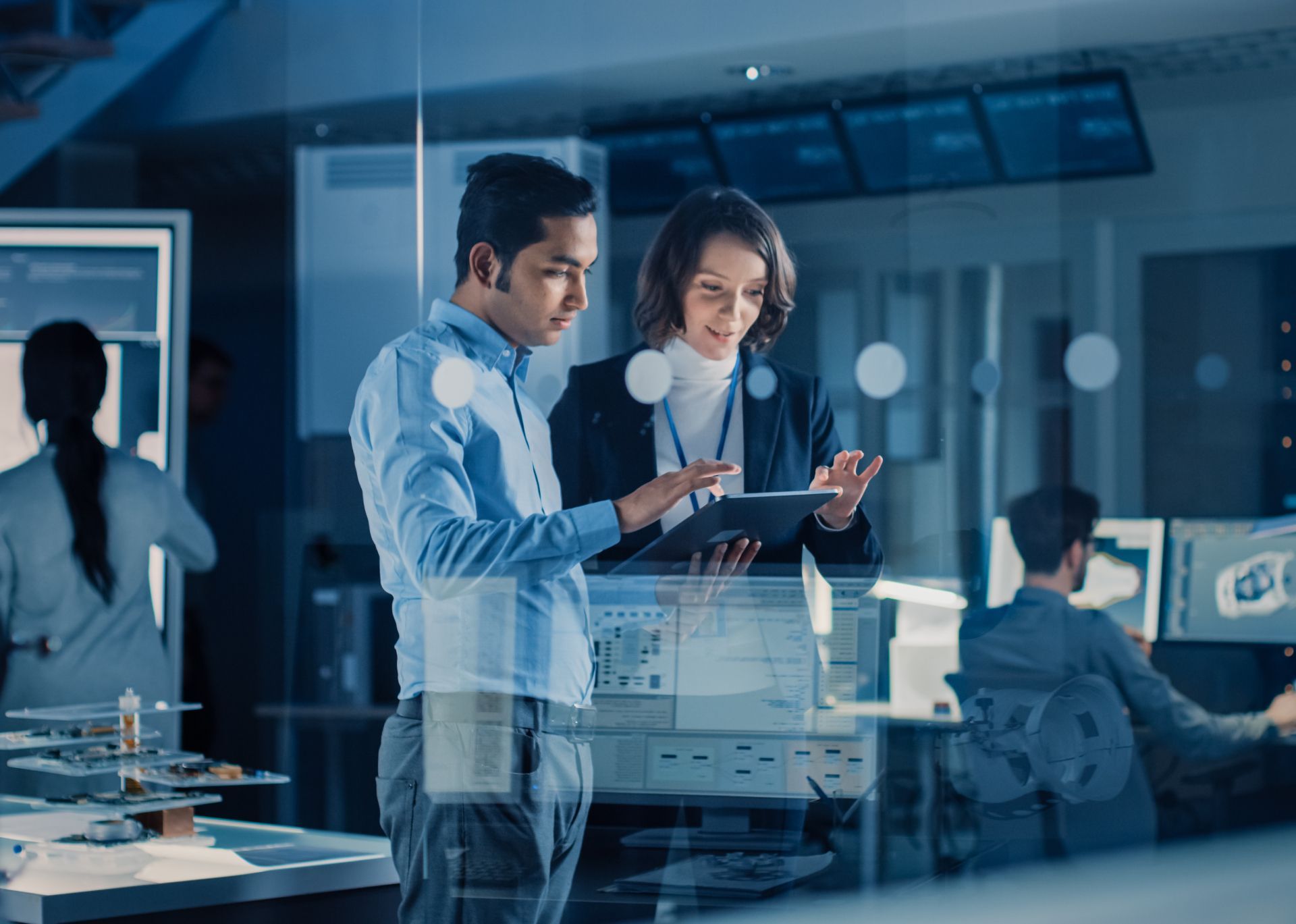 Two professionals reviewing data on a tablet in a modern glass-walled office with screens in the background