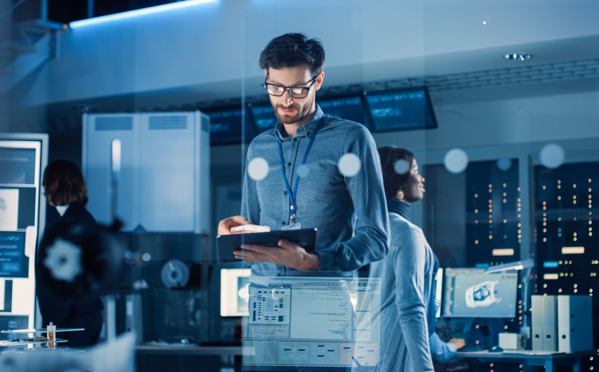 Technician using a tablet in a blue-lit server room with computer monitors and data center equipment