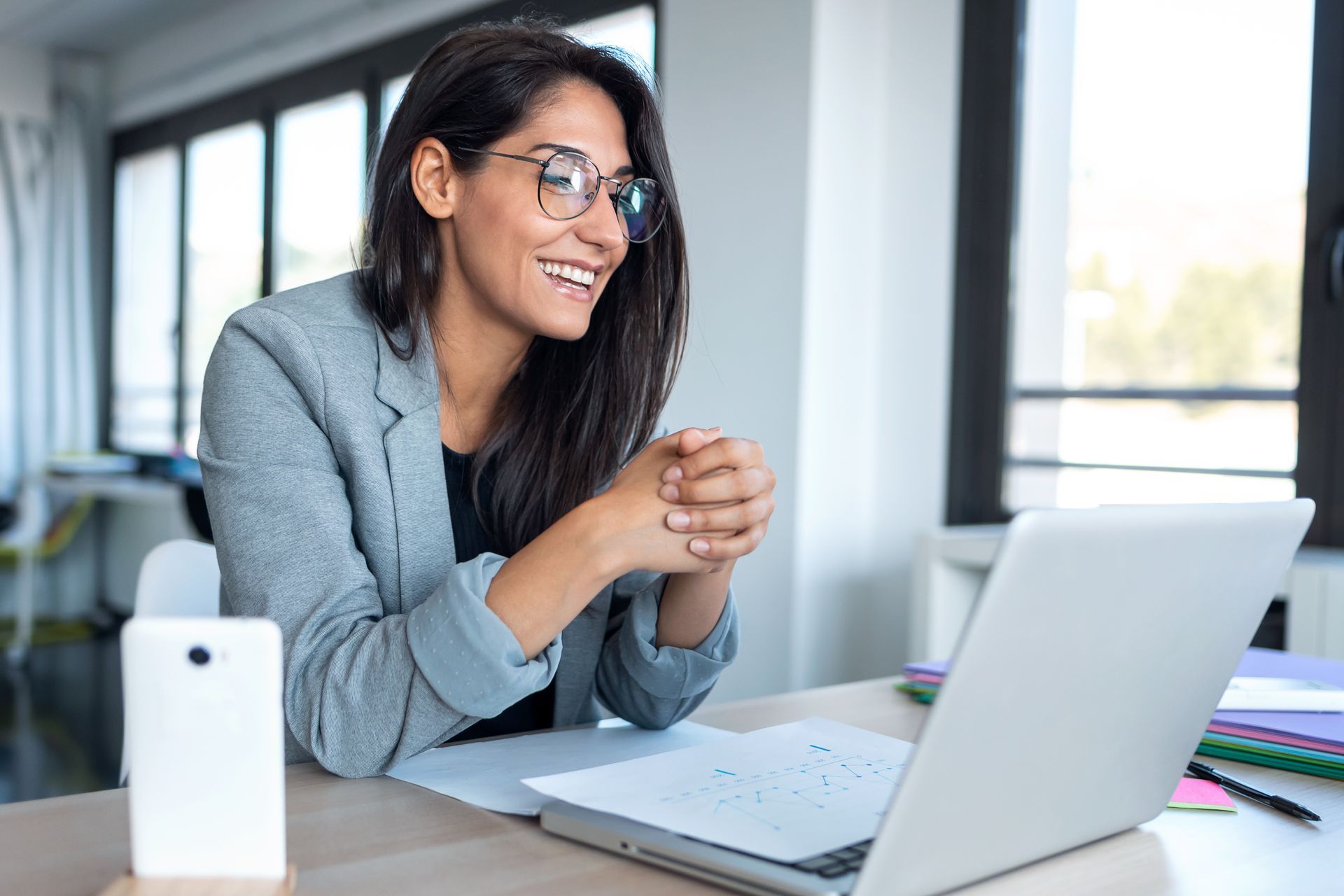 Woman in a gray blazer smiles at a desk, working on a laptop in a bright office
