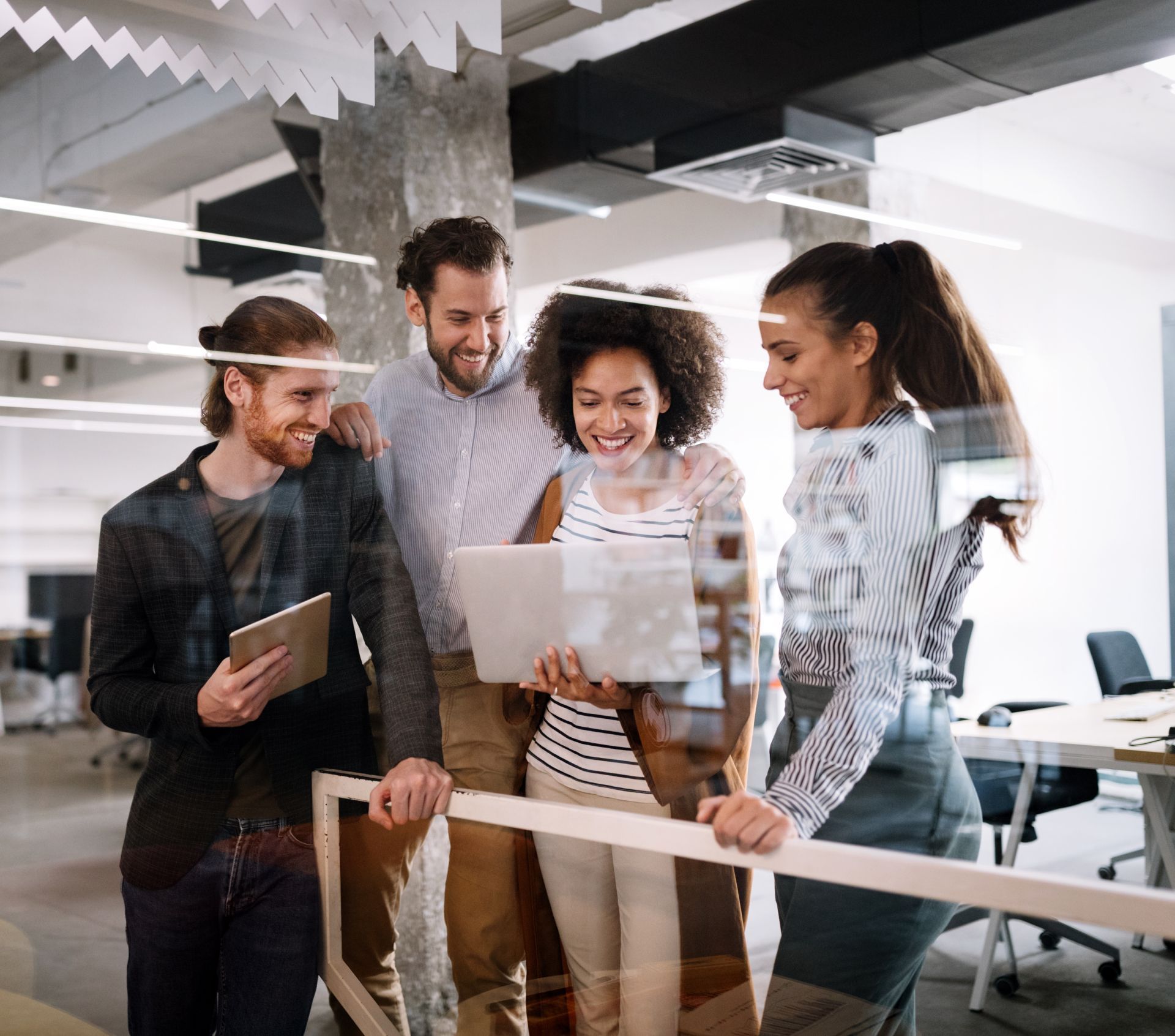 Four coworkers smiling and collaborating around a laptop in a bright office space