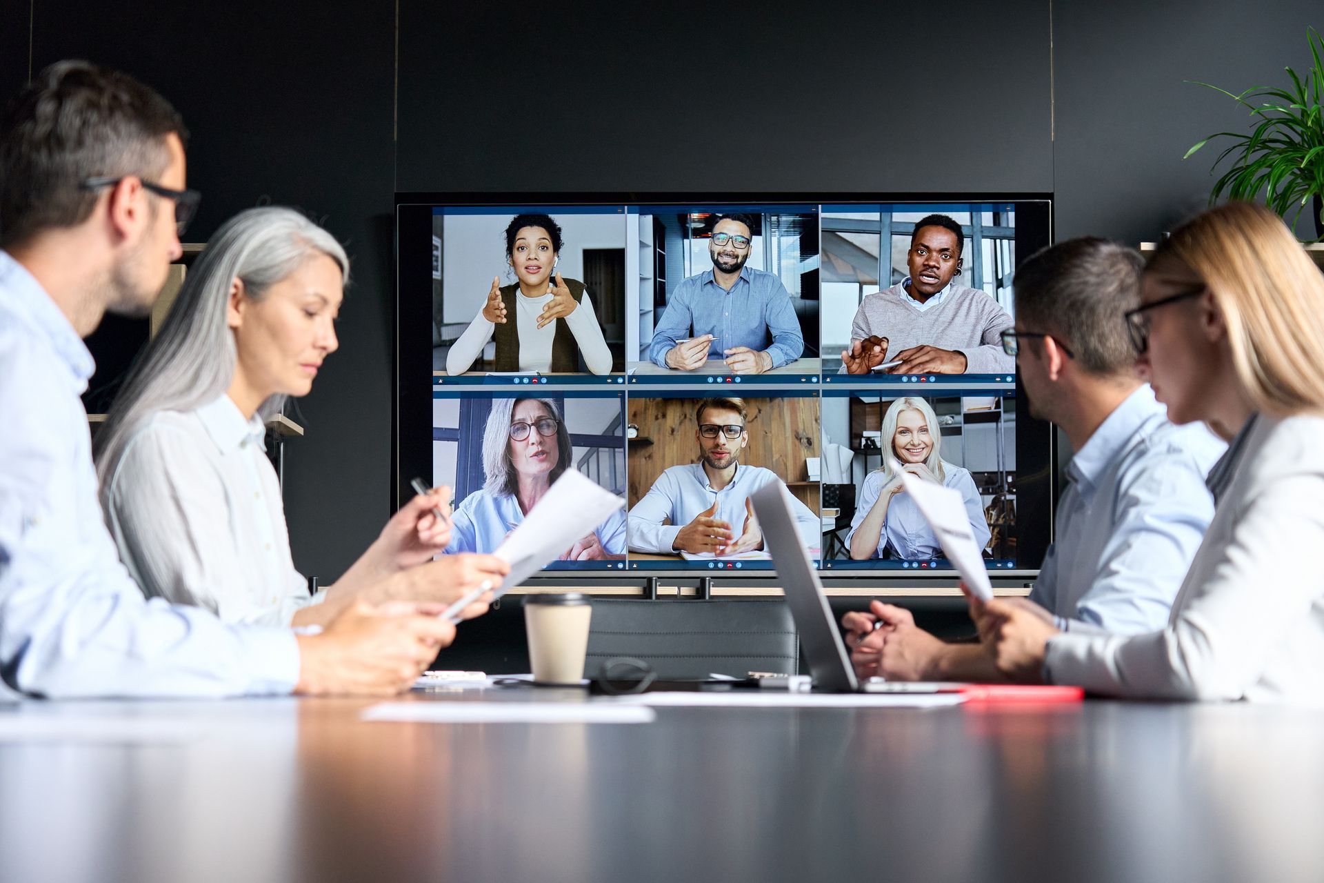 Business meeting with six people in a conference room, video conferencing with a remote participant on a large screen
