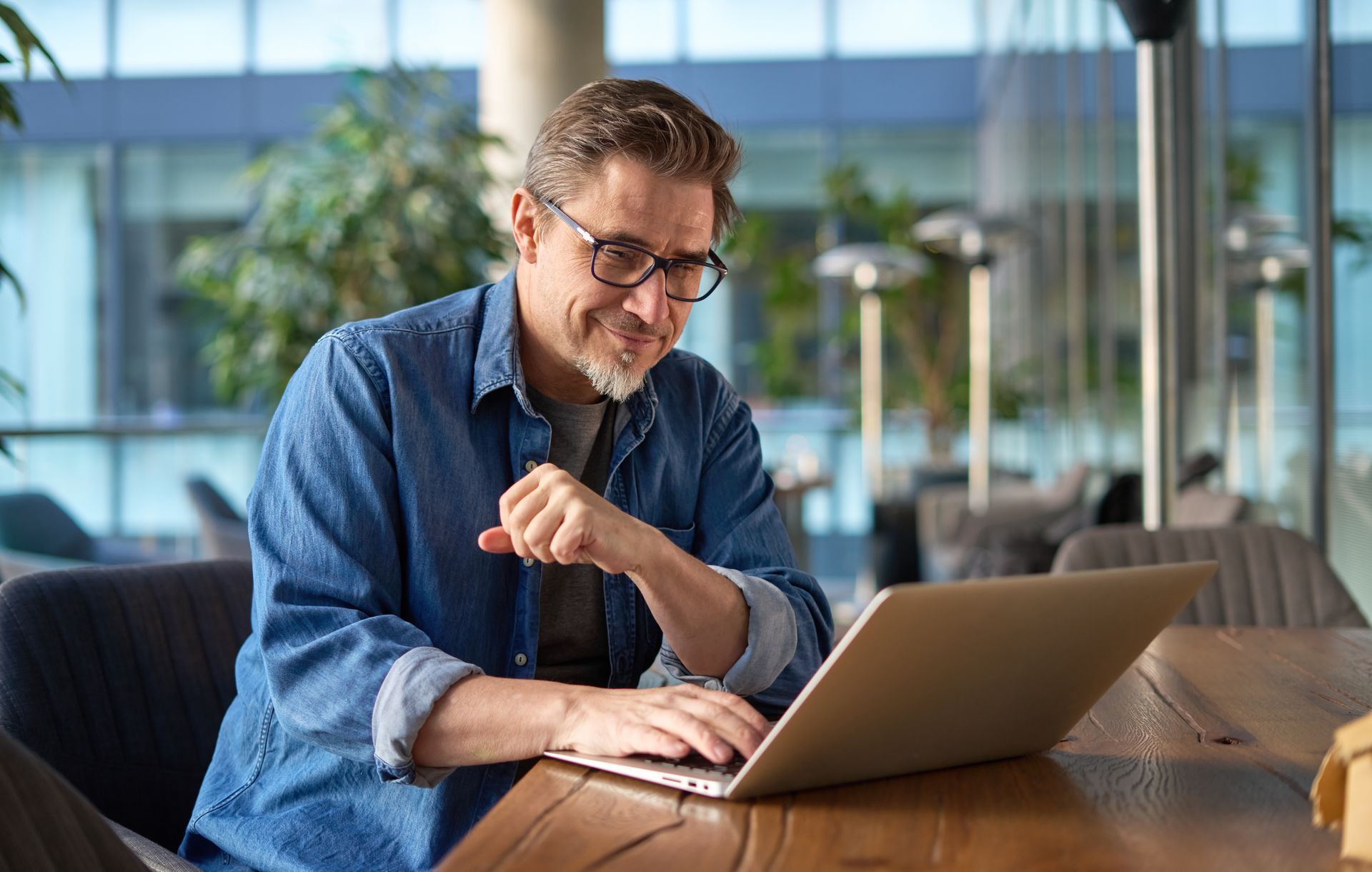 Man in glasses using a laptop at a wooden table in a bright office lounge