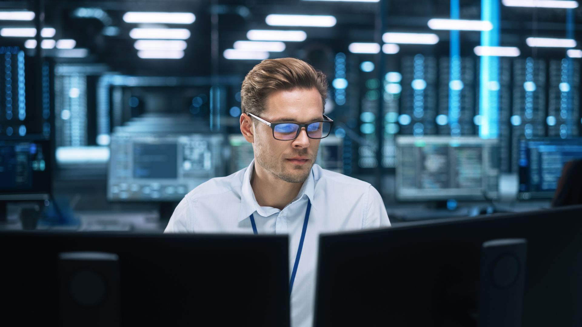 Man working at a computer in a modern server room, wearing glasses and a white shirt.