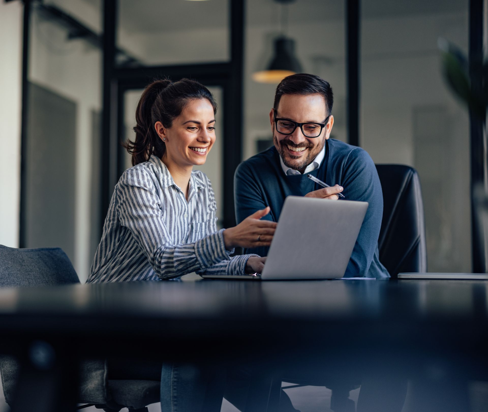 Two coworkers smiling and reviewing a laptop in a glass-walled office