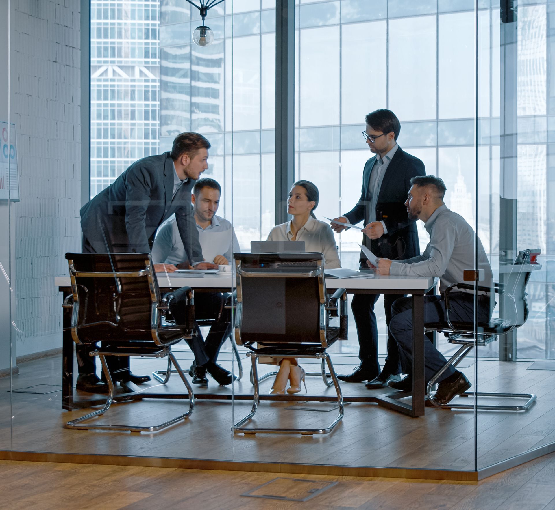 Team meeting in a glass-walled conference room, with five professionals discussing documents around a table.