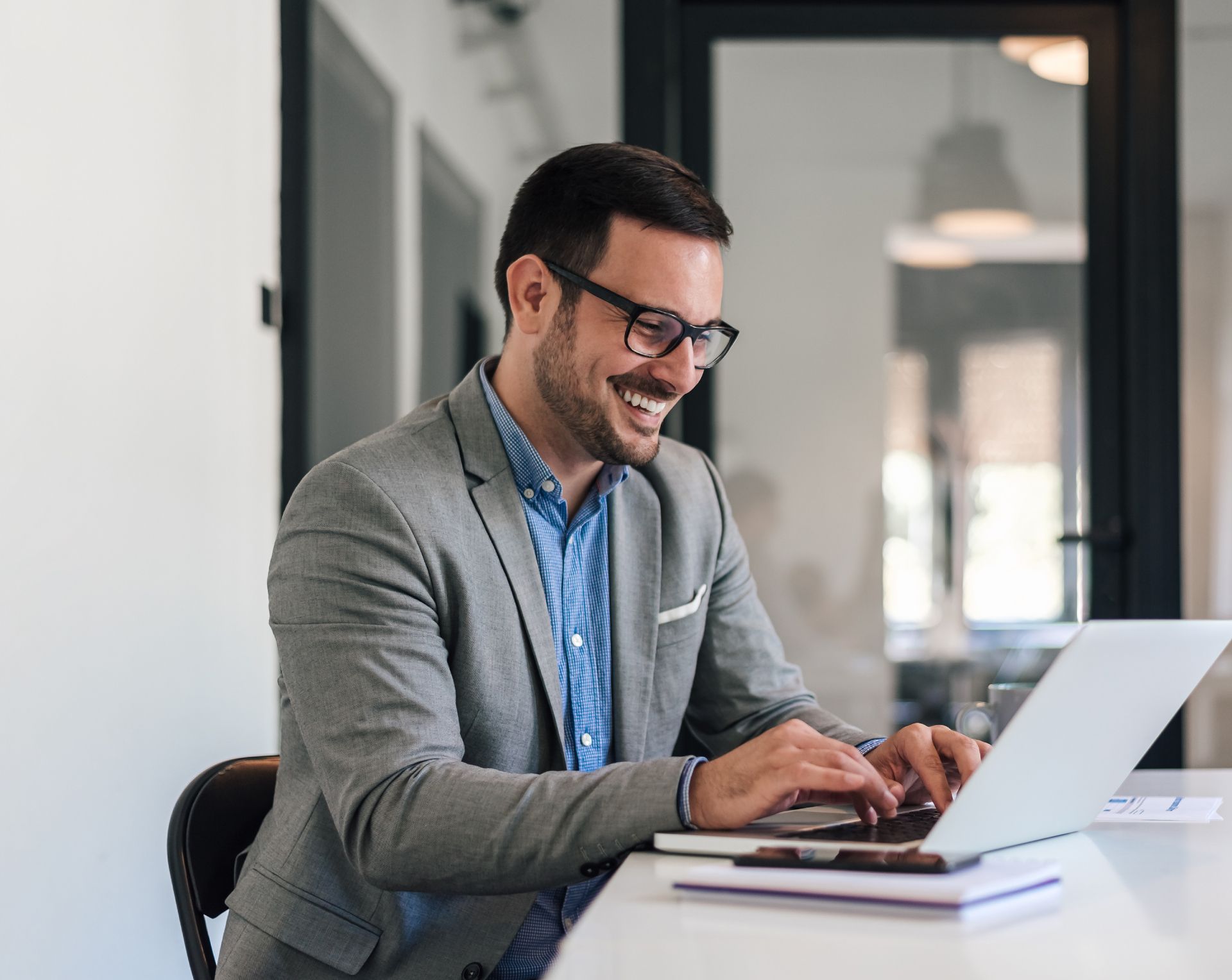 Man in glasses typing on a laptop at a desk in a bright office