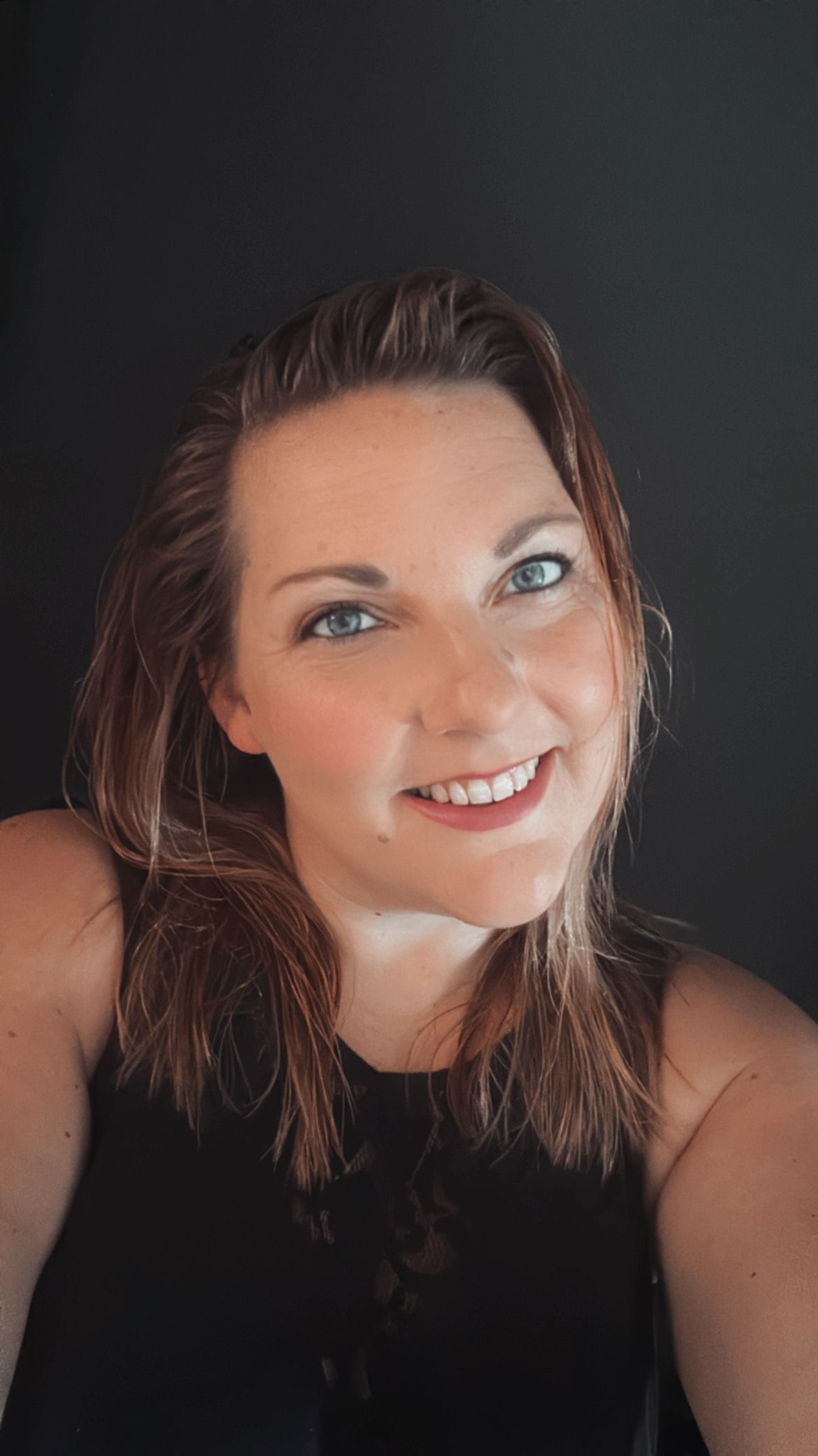 Lachende vrouw met golvend bruin haar in een zwarte top tegen een donkere achtergrond.