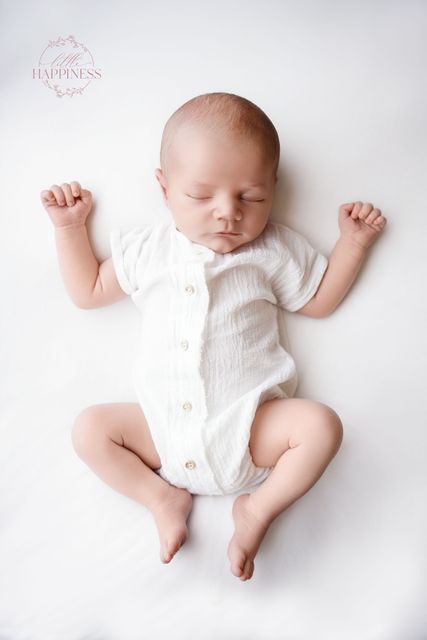 Slapende baby in een wit rompertje op een witte achtergrond, armen gebogen en benen ontspannen.