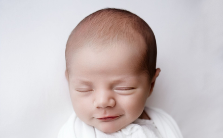 Slapende baby met gesloten ogen, gewikkeld in een witte deken, op een lichte achtergrond.