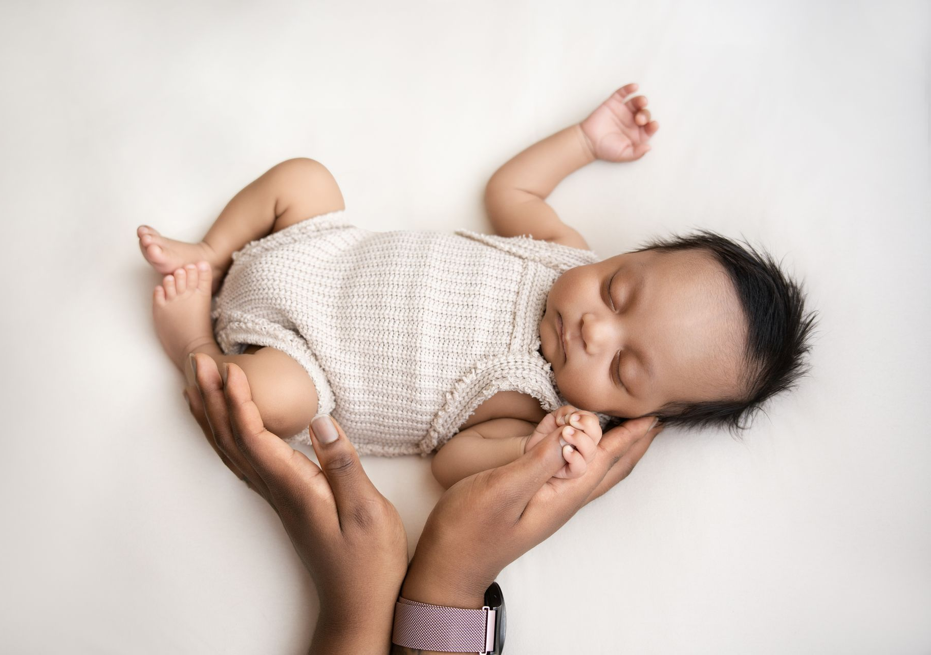 Slapende baby in een wit gebreid pakje, gewiegd in de handen van een volwassene, op een witte achtergrond.